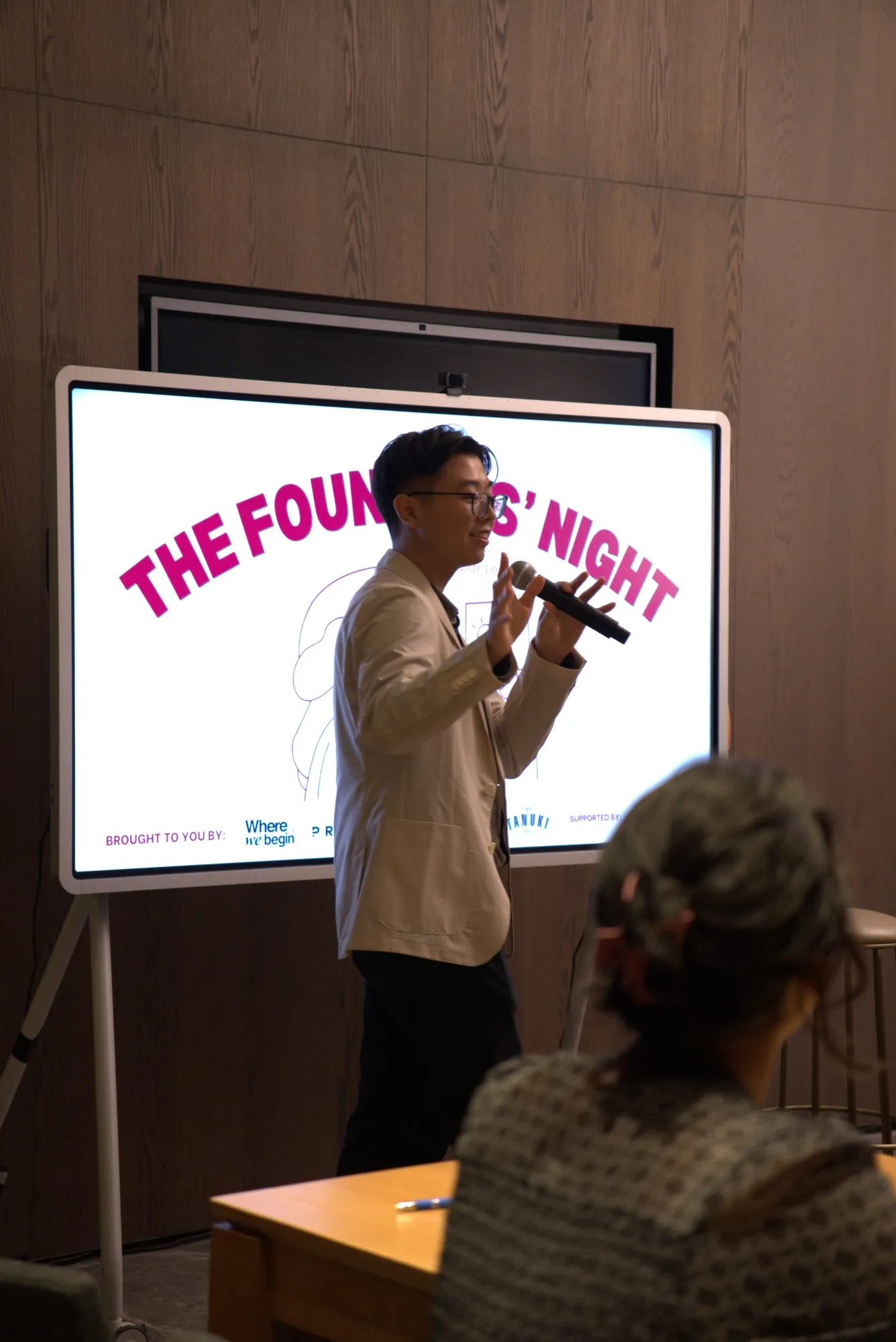 Benedict speaking into a microphone in front of a screen reading “The Founders’ Night”, with an audience member in the foreground.