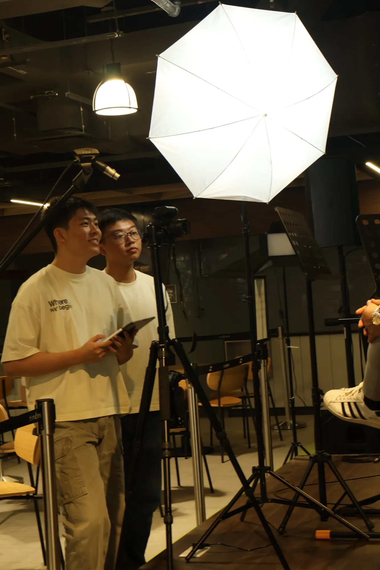 Benedict and a teammate in WhereWeBegin T-shirts standing behind a camera and an umbrella light, watching an interview being filmed.