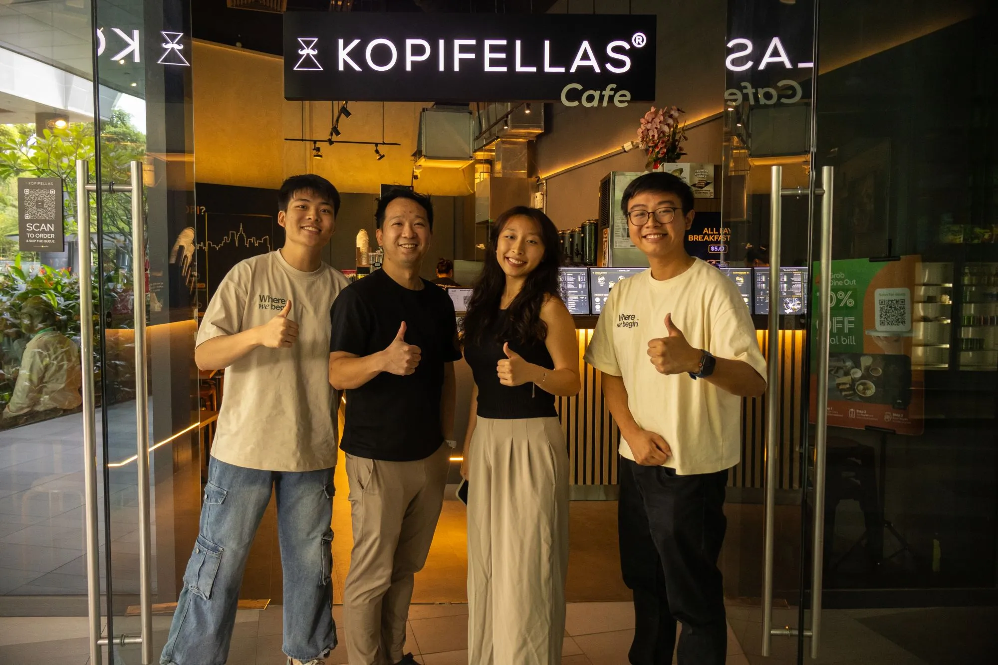 Benedict and three others giving a thumbs-up at the entrance of Kopifellas Cafe, two of them in WhereWeBegin T-shirts.