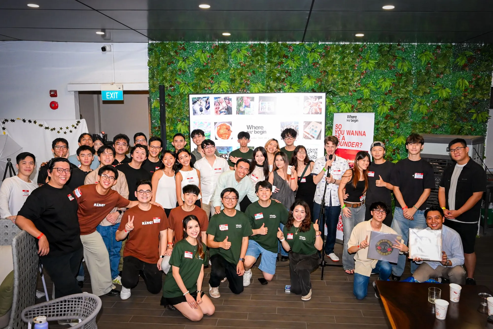 A large group of founders and attendees posing together at a WhereWeBegin event, in front of a photo wall and a banner reading “So you wanna be a founder?”