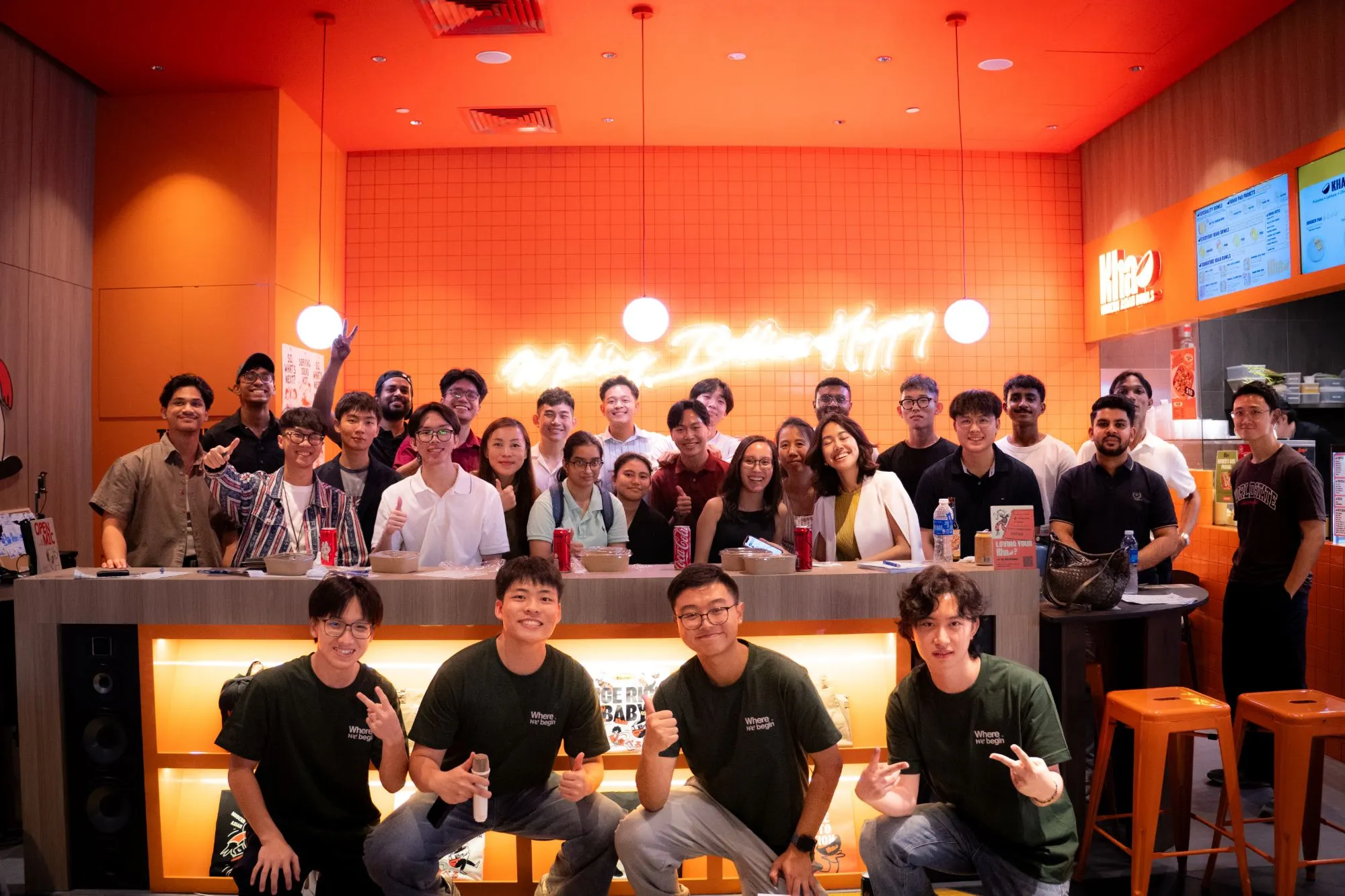 Around thirty attendees and four WhereWeBegin team members in green shirts posing at an orange-tiled restaurant counter under a neon sign.